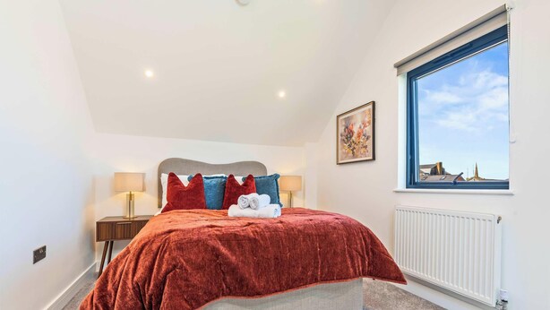 A bedroom with a bed, two bedside lamps, a radiator, and a window with a city view. - New Central Home | Groups, Contractor & Families (Reading)