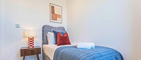 Uma cama bem arrumada com manta e almofadas azuis, uma luminária de cabeceira e uma pequena mesa de cabeceira de madeira.
