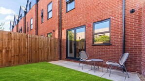 A brick building with a wooden fence, a patio with chairs, and a green lawn. - New Central Home | Groups, Contractor & Families (Reading)