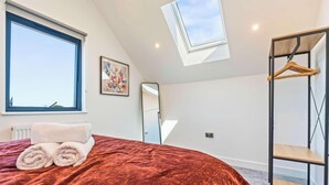 A bedroom with a large window, a skylight, a bed with rolled towels, a wooden hanger, and a radiator. - Modern Reading House|Contractor & Family Friendly (Reading)