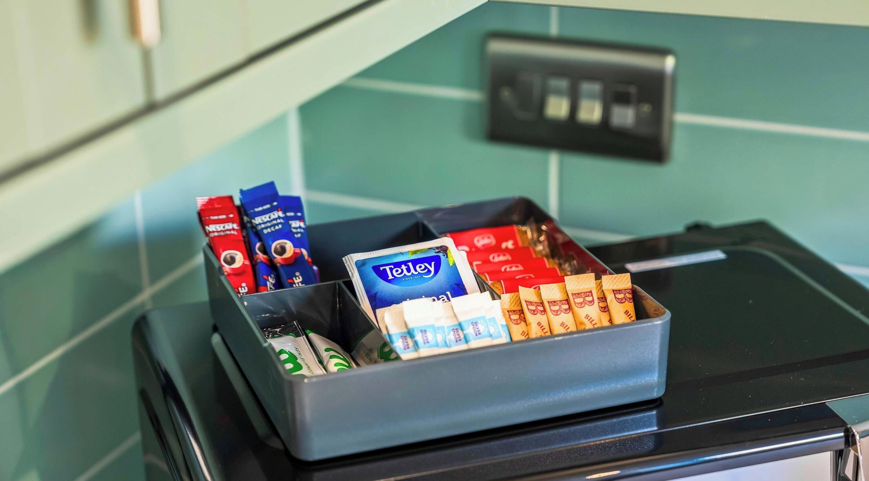 Ein Tablett mit verschiedenen verpackten Lebensmitteln, darunter eine blaue Tetley-Tee-Verpackung, rote und blaue Nestle Original Entkoffeiniert-Verpackungen und mehrere Schokoladentafeln.