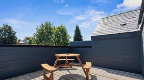 Eine Dachterrasse mit einem Holztisch und Bänken, umgeben von einer dunkel gefärbten Mauer und einem geneigten Dach.