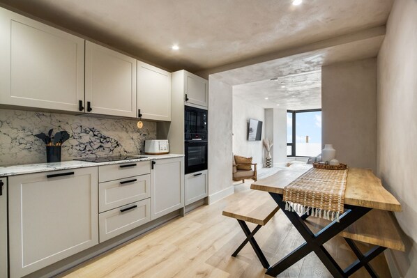 Een moderne keuken met witte kastjes, een marmeren spatwand, en een eethoek met een houten tafel en bank.