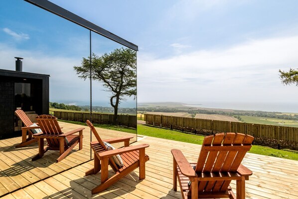 A deck with wooden chairs and a table, overlooking a rural landscape with fields and a distant view of the sea.