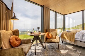 A modern hotel room with a large window, a sofa, a chair, a small table with drinks, and a bed.