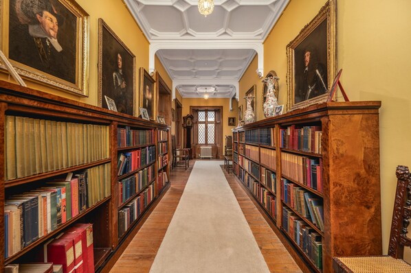 A library with bookshelves on both sides, a central carpeted walkway, and a ceiling with decorative elements.