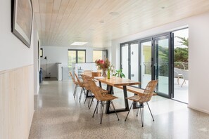 A modern dining area with a wooden table and wicker chairs, large windows, and a view of greenery outside.