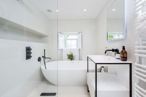 A modern bathroom with a freestanding bathtub, a white vanity with a mirror, and a window with shutters.