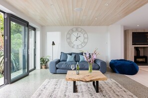 A modern living room with a large window, a blue sofa, a wooden coffee table, and a wall clock.