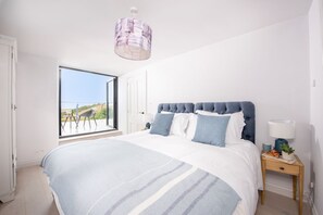 A bedroom with a large bed, a view through an open door, a bedside table with a lamp, and a hanging light fixture.