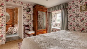 A bedroom with floral wallpaper, a large bed, a wooden wardrobe, a mirror, and a window with curtains.