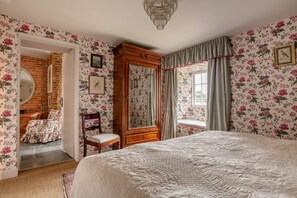 A bedroom with floral wallpaper, a large bed, a wooden wardrobe, a mirror, and a window with curtains.