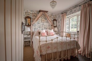 A bedroom with a four-poster bed, a nightstand with a lamp, a window with curtains, and a chandelier.