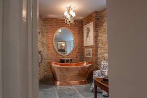 A bathroom with a copper bathtub, a round mirror, a chandelier, and a brick wall.
