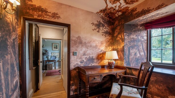 A room with a wooden desk, a chair, a window with a view of greenery, and a door leading to another room.