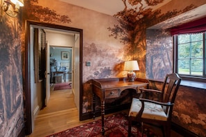A room with a wooden desk, a chair, a window with a view of greenery, and a door leading to another room.