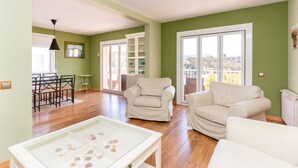 A spacious living room with a glass coffee table, two beige armchairs, a dining area with a table and chairs, and a view through a sliding glass door. - Apartment Limoncello (Sant Pere de Ribes)