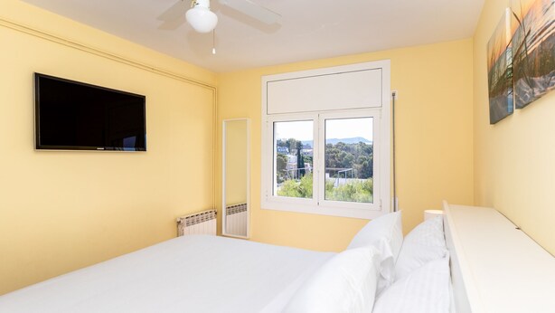 A bedroom with a bed, a television, a window with a view, and a ceiling fan. - Apartment Limoncello (Sant Pere de Ribes)