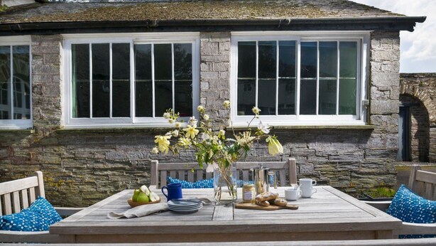 Un bâtiment en pierre avec une table en bois dressée pour le petit-déjeuner, agrémentée d’un vase de fleurs, d’assiettes, de tasses et d’une cafetière.
