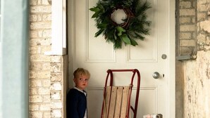 Un enfant se tient devant une porte, avec une couronne de Noël, une échelle rouge et un cadeau emballé.