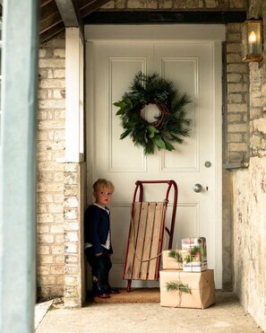 A child standing at a door with a wreath, a red ladder, and a wrapped gift.