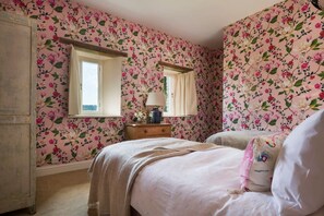 A bedroom with floral wallpaper, a wooden dresser, a bedside table with a lamp, and a bed with a white pillow.
