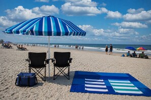 A beach with two black folding chairs, a blue beach mat, and a striped beach umbrella.