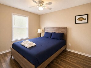 A bedroom with a bed, a ceiling fan, a window with blinds, a bedside lamp, and a framed picture on the wall.
