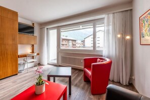 Una habitación moderna con un armario de madera, un sofá rojo, una mesita y vistas a edificios a través de amplios ventanales.