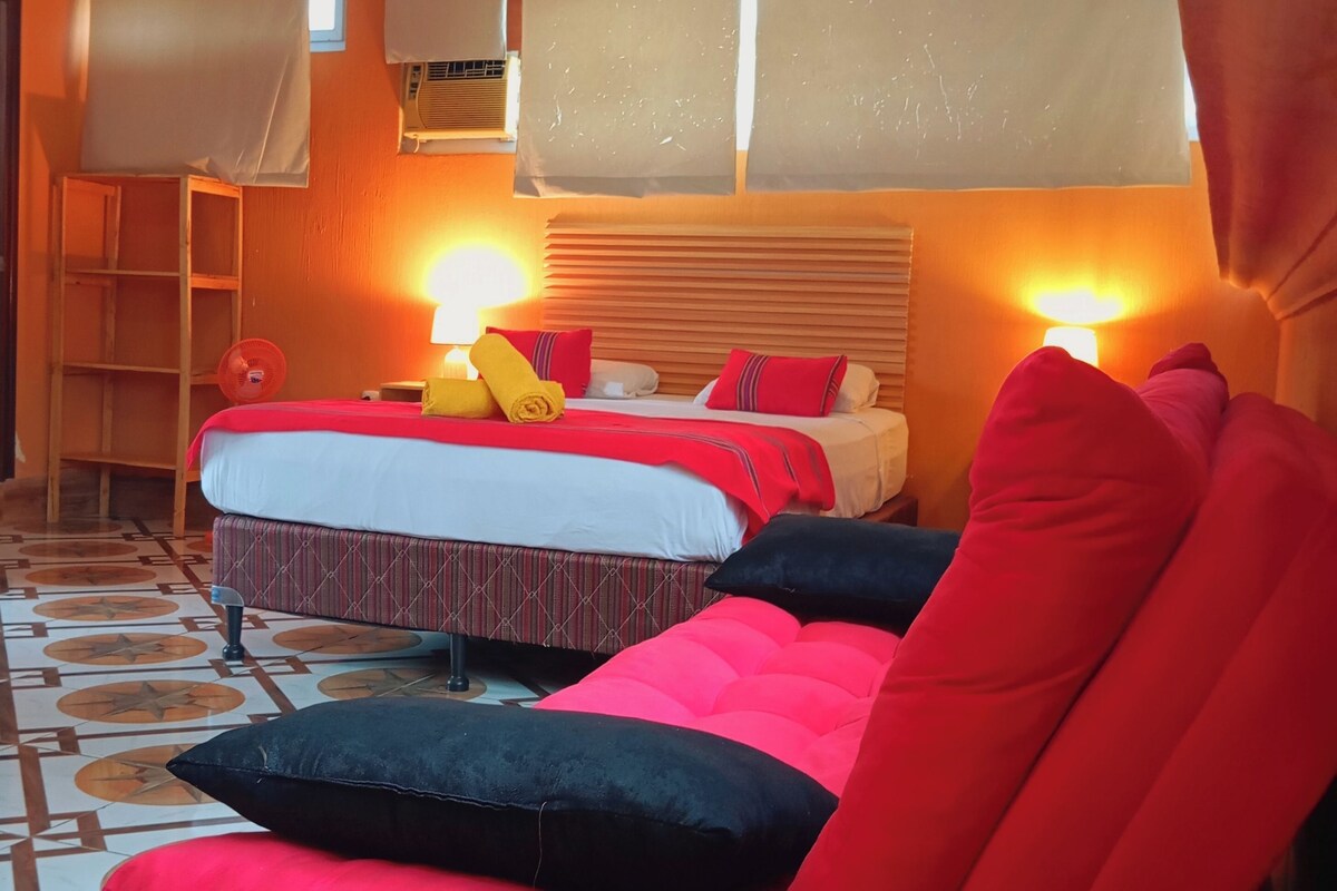 Habitación de hotel con dos camas, una silla roja, un ventilador de pie y un ventanal con cortinas.
