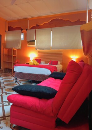 Habitación de hotel con dos camas, una silla roja, un ventilador de pie y un ventanal con cortinas.