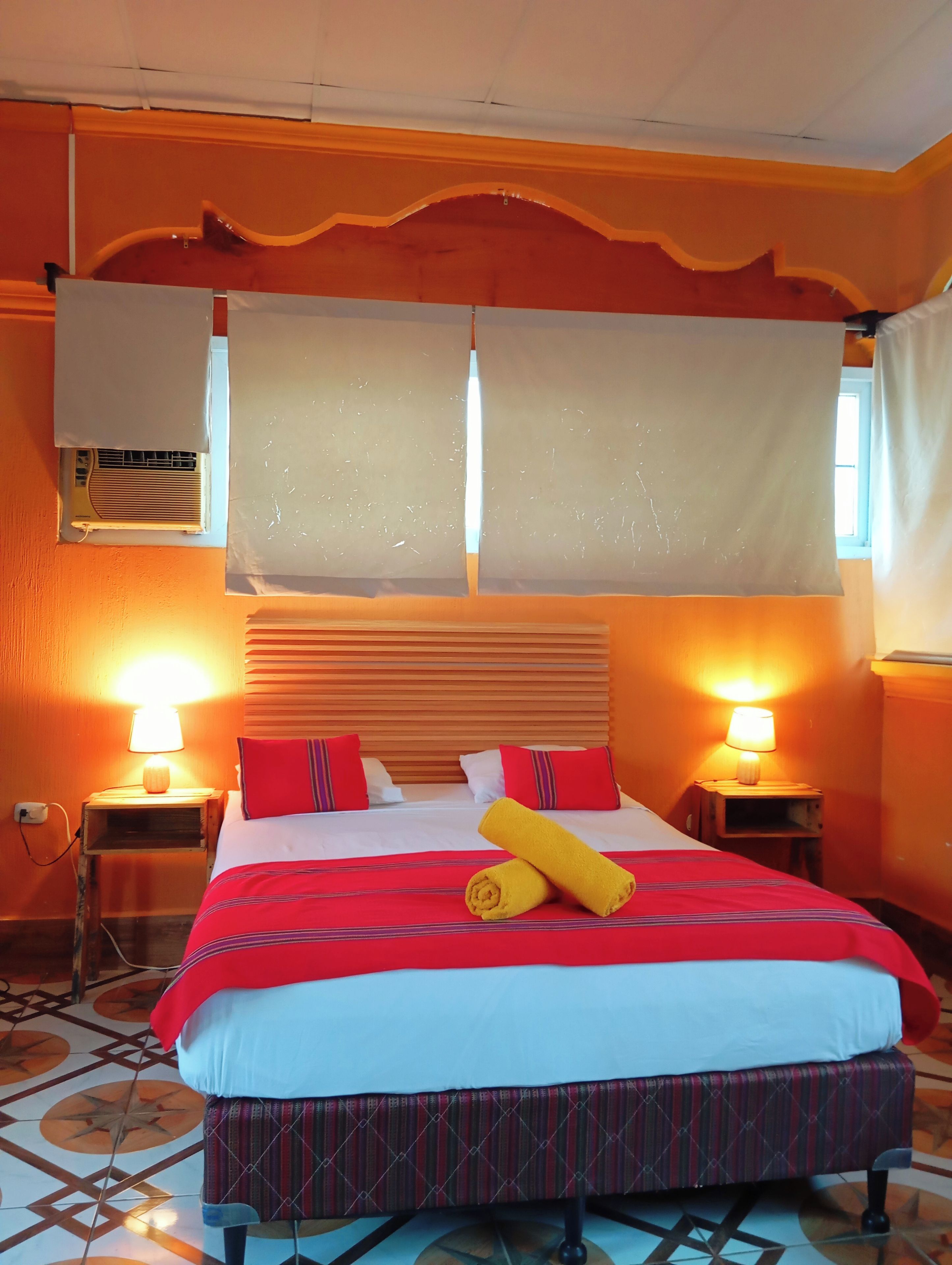 Una habitación de hotel con una cama, dos lámparas de noche, un ventanal con cortinas y un piso de baldosas.