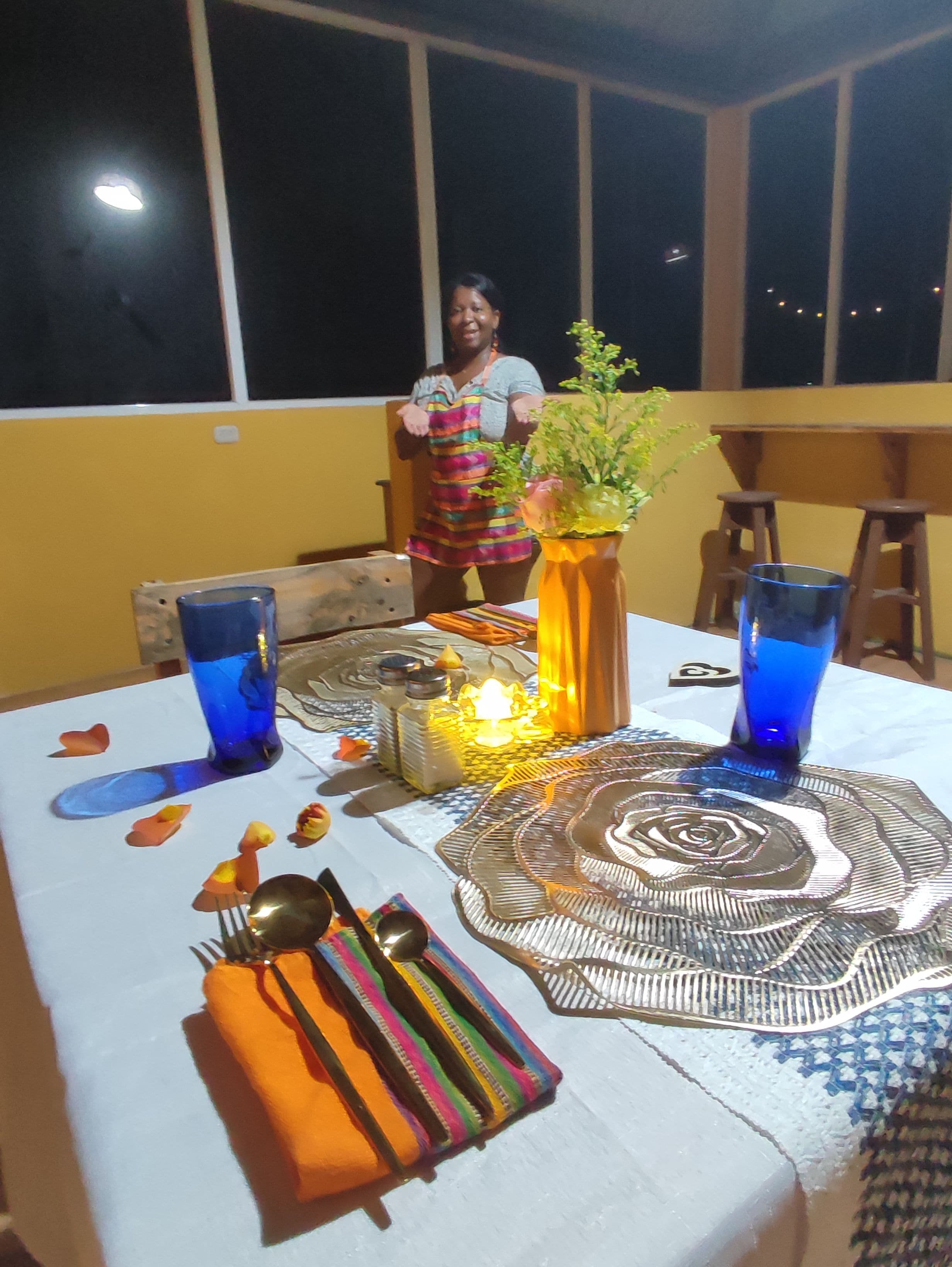 Mesa de jantar posta com copos azuis, guardanapo colorido e um centro de mesa decorativo.