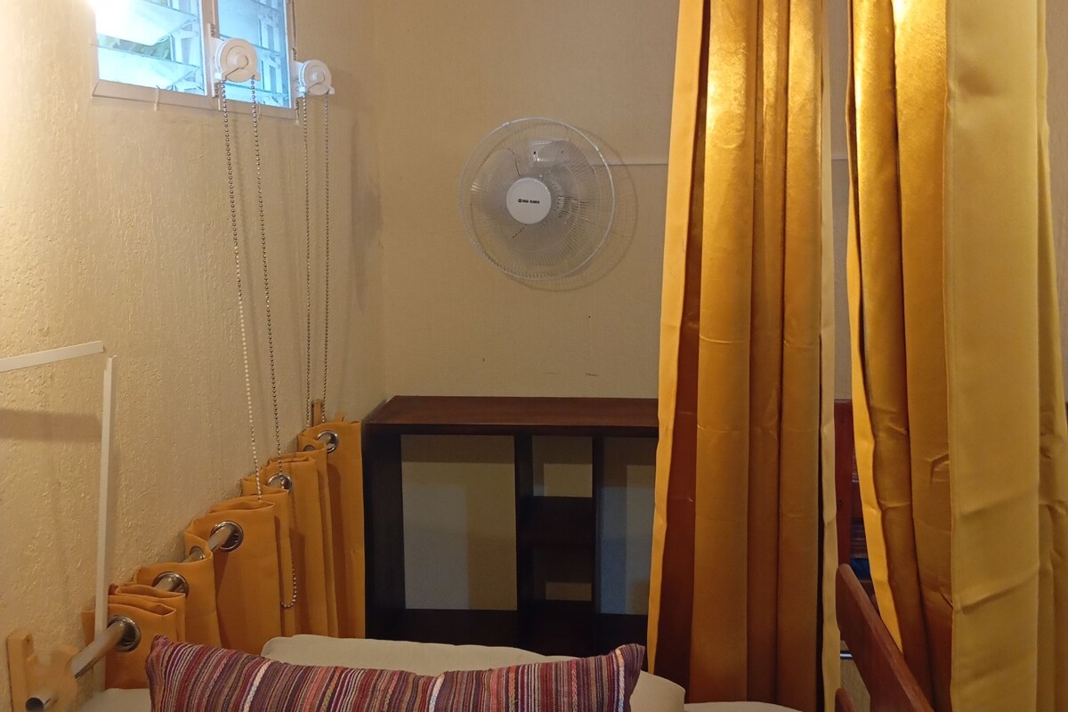 Habitació d'hotel amb un llit, un ventilador de peu, cortines i una finestra amb persianes.