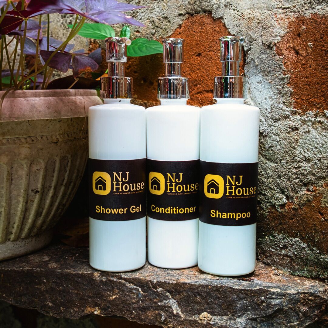 Tres botellas de productos NJ House: Gel de ducha, acondicionador y champú, sobre un repisa de piedra con una planta en maceta al fondo.