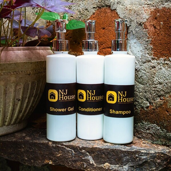 Tres botellas de productos NJ House: Gel de ducha, acondicionador y champú, sobre un repisa de piedra con una planta en maceta al fondo.