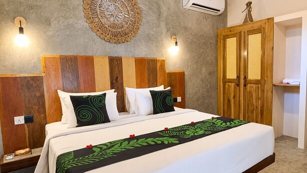 A hotel room with a wooden headboard, a bed with white bedding and green decorative pillows, a wall-mounted air conditioner, and a wooden wardrobe with a woven door. - Private Pool Villa (Tangalle)