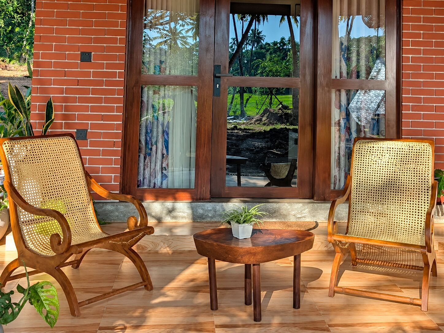 Une façade en briques, une porte et des fenêtres en verre, deux fauteuils en osier, une petite table ronde et une plante en pot.