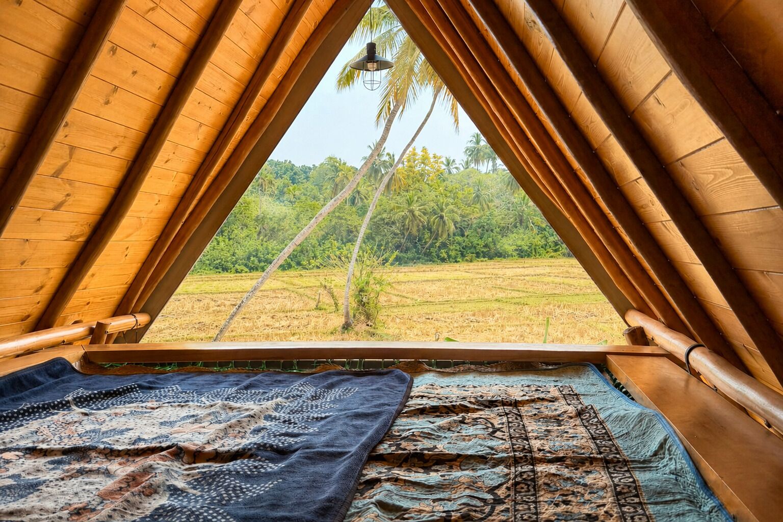 A room with a sloped wooden ceiling, a bed with patterned bedding, and a view of a palm tree and fields.