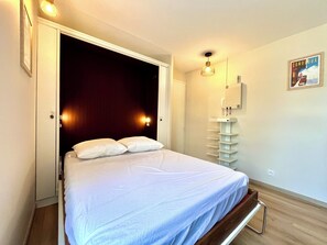 A bedroom with a bed, a bedside table, a wall-mounted shelf, and a poster on the wall. - Studio in the heart of Combloux near the lake (Combloux)