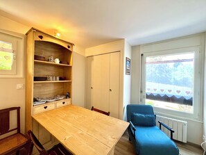 A room with a wooden table, a blue chair, a wardrobe, and a window with a view of trees. - Studio in the heart of Combloux near the lake (Combloux)