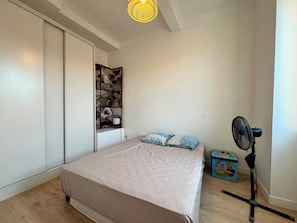 A bedroom with a bed, a wall-mounted shelf with decorative items, a standing fan, and a small toy chest. - Newly renovated apart in the heart of Old Antibes (Antibes)