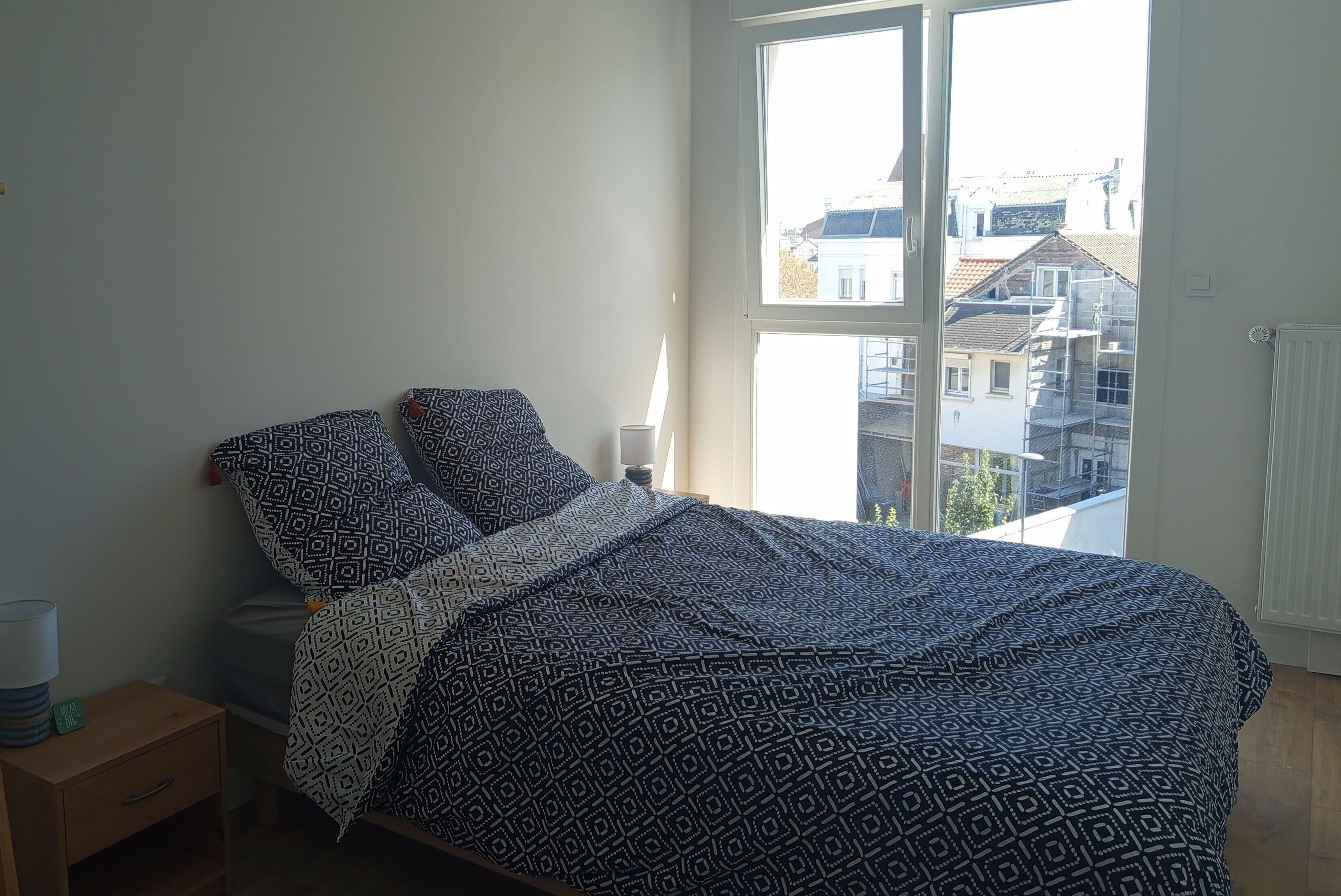 A bedroom with a bed, a nightstand, a lamp, and a window with a view of buildings.