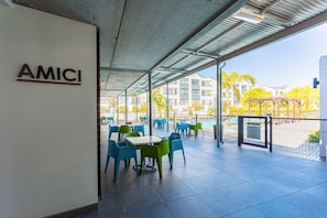 An outdoor patio area with tables and chairs, a sign reading "AMICI", and a view of buildings and trees in the background. - Modern 3-bed Garden Pool Gym by Ctha (Cape Town)