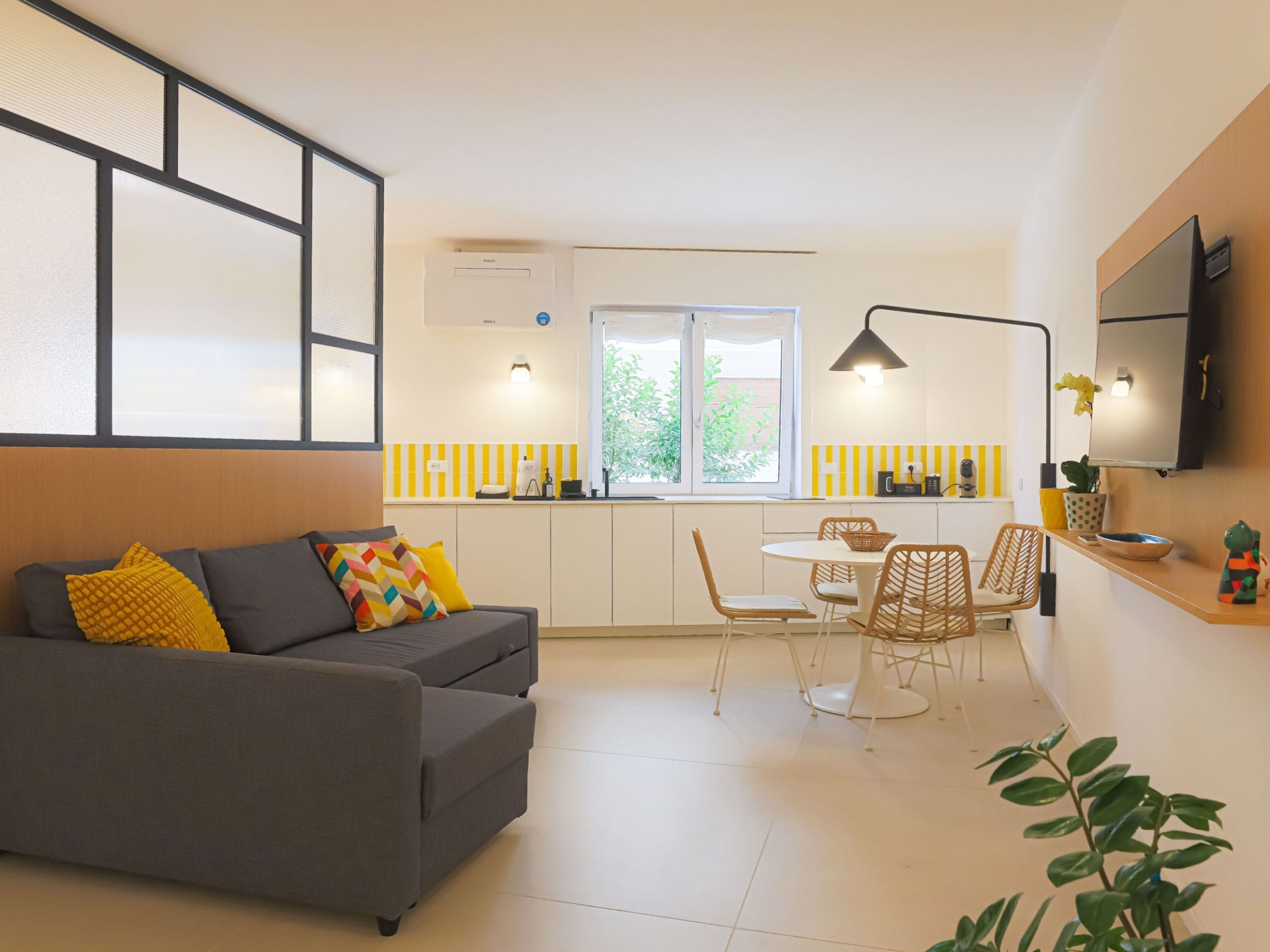 Una sala de estar moderna con sofá, una mesa de comedor con sillas, una zona de cocina con ventana y un televisor de pantalla plana montado en la pared.