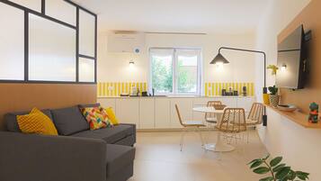 Una sala de estar moderna con sofá, una mesa de comedor con sillas, una zona de cocina con ventana y un televisor de pantalla plana montado en la pared.