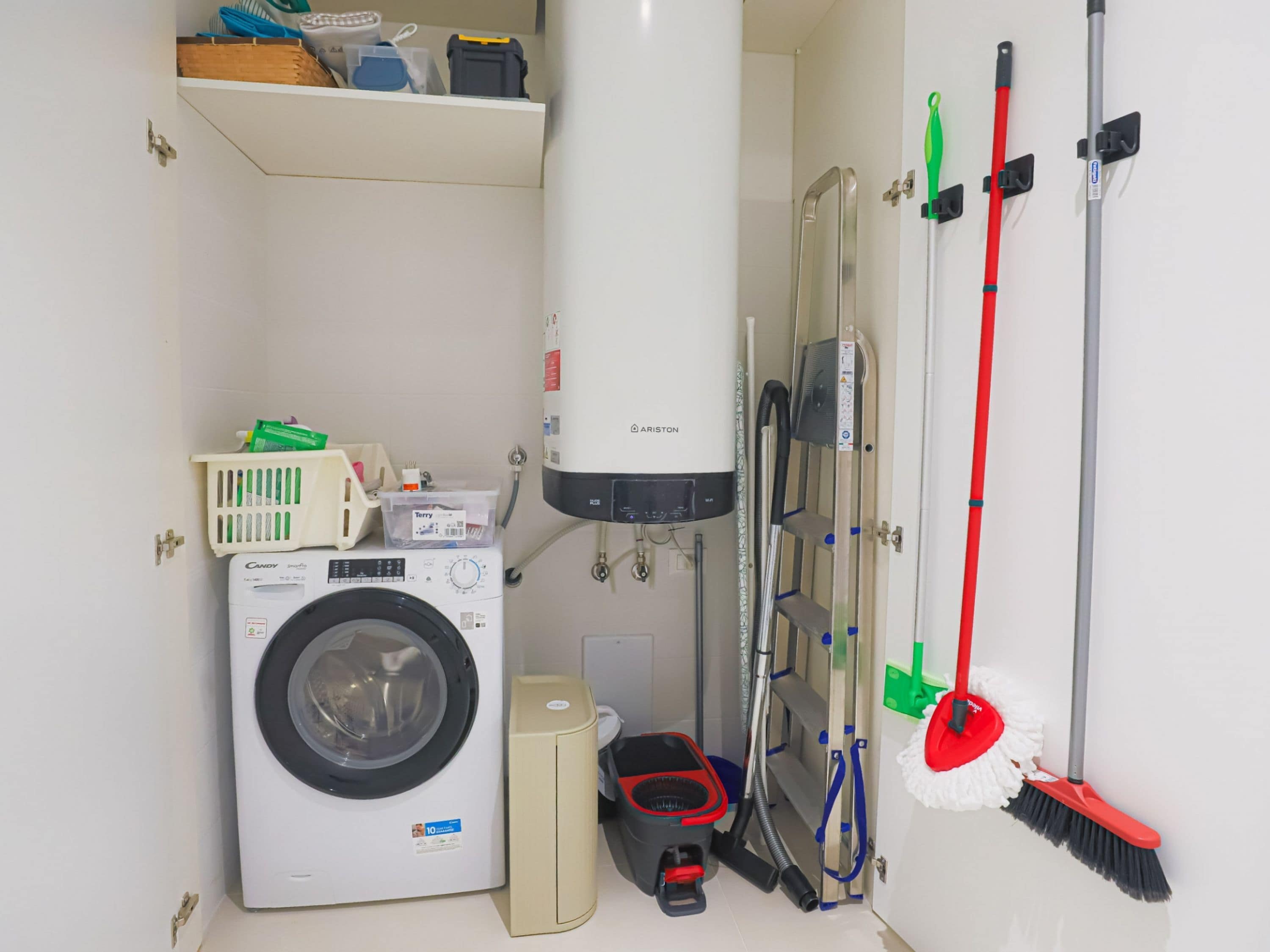 A compact laundry room with a front-loading washing machine, a tall water heater, a vacuum cleaner, and a mop rack.