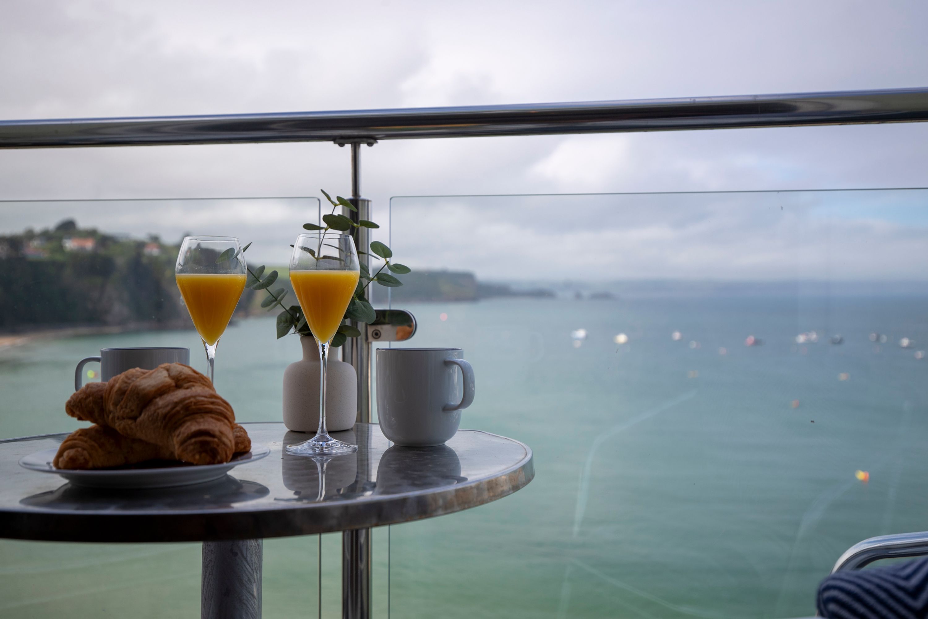 Ein Tisch mit Croissants, einem Orangensaftglas und einer Tasse, mit Blick auf ein Gewässer mit Booten.