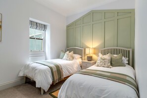 A bedroom with two beds, a window with a striped valance, and a green headboard. - Royal Victoria Court 7 - Tenby Harbour Sea Views (Tenby)