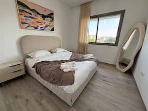 A modern hotel room with a bed, a bedside table, a large mirror, and a window with a view of buildings. - Malaga 2 Bright Luxury 1BR Central Location (La Marsa)